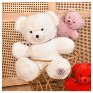 White plush teddy bear with a striped ribbon sits in a wicker chair, with a pink teddy bear and a red teddy bear nearby.