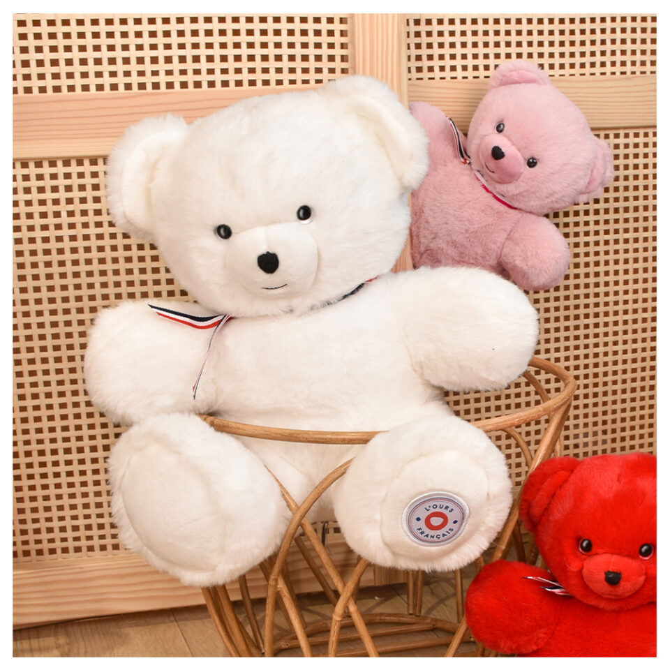 White plush teddy bear with a striped ribbon sits in a wicker chair, with a pink teddy bear and a red teddy bear nearby.