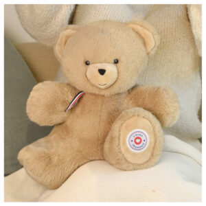 Soft beige teddy bear plush with a red, white, and blue ribbon, sitting on a white blanket or cushion.