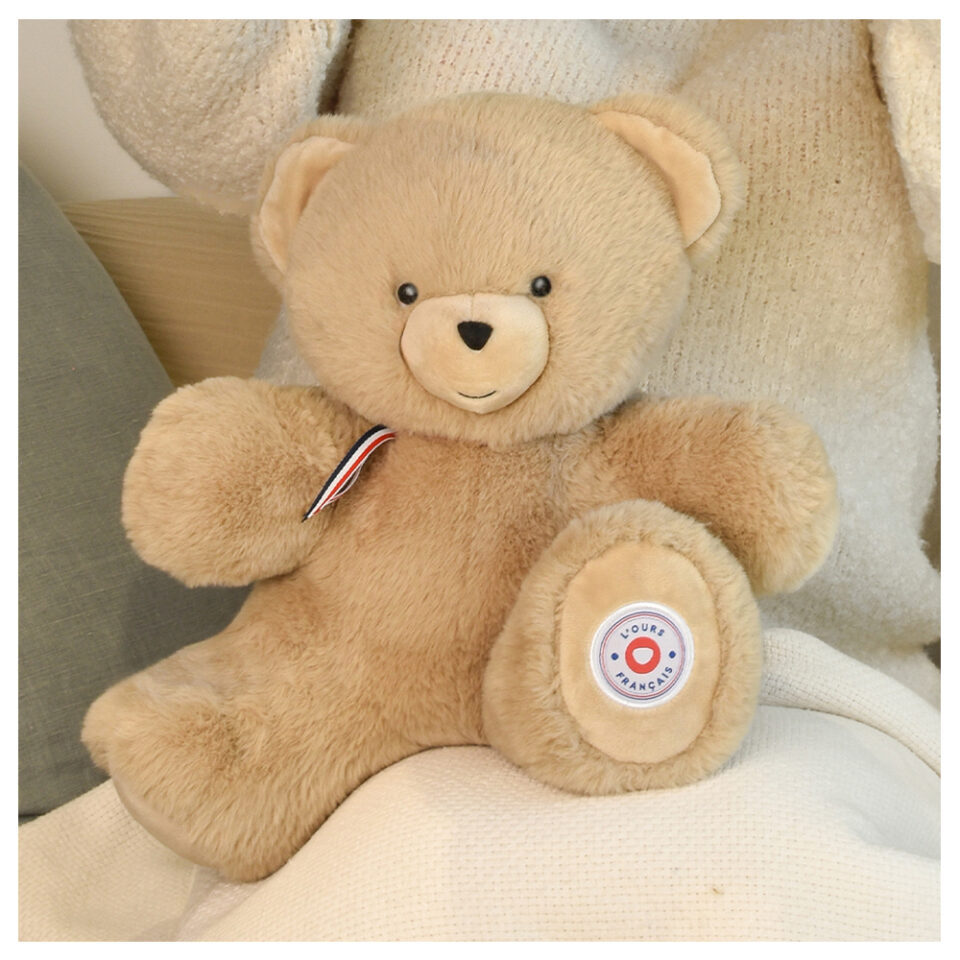 Soft beige teddy bear plush with a red, white, and blue ribbon, sitting on a white blanket or cushion.