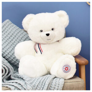 White plush teddy bear with a red, white, and blue ribbon around its neck sits on a beige armchair with a gray knitted blanket draped over it.