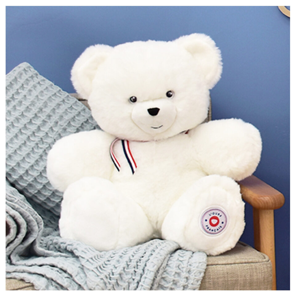 White plush teddy bear with a red, white, and blue ribbon around its neck sits on a beige armchair with a gray knitted blanket draped over it.