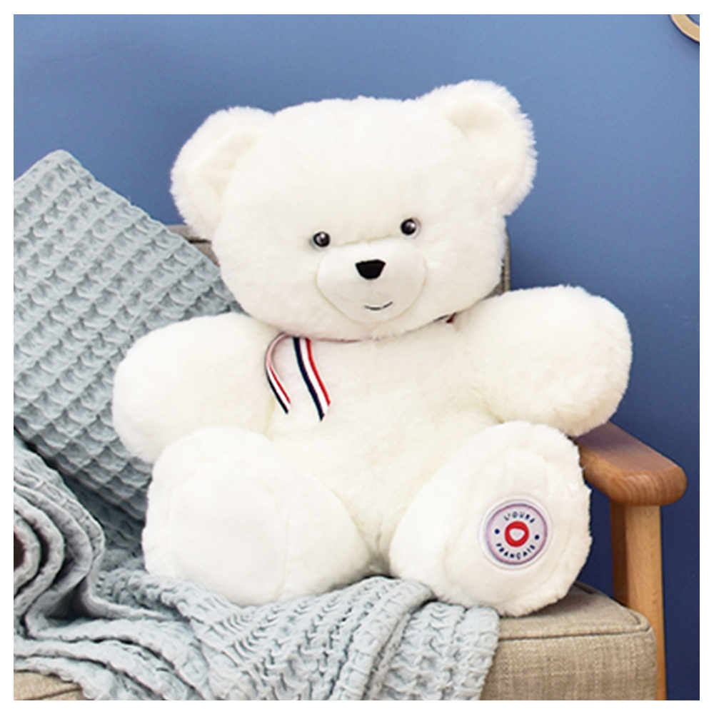 FR8052-ambiance – L'Ours Français White plush teddy bear with a red, white, and blue ribbon around its neck sits on a beige armchair with a gray knitted blanket draped over it.